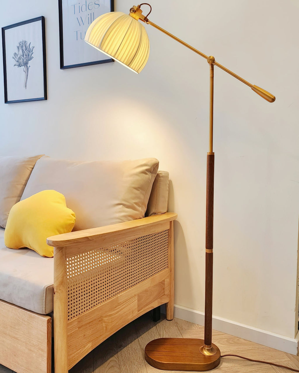 Close crop of the Clérachic floor lamp's lit shade, wooden stem and oval base beside a rattan-sided sofa with a yellow cushion.