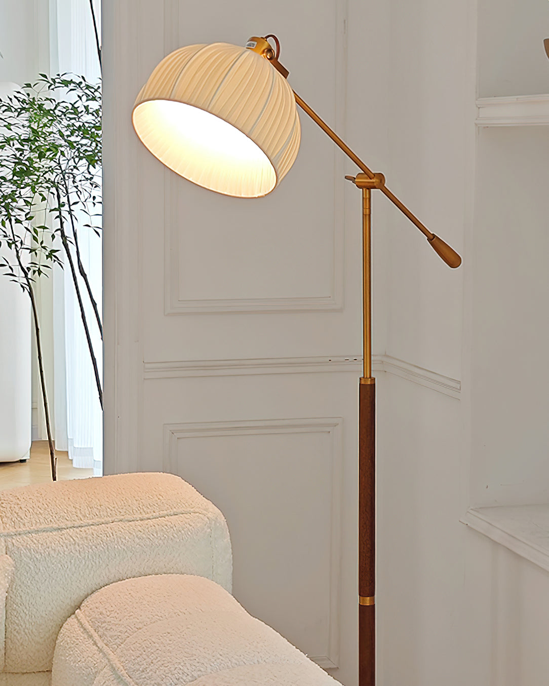 Close view of the pleated beige dome shade and brass arm of this Clérachic floor lamp, with a white panelled door and a cream seat.