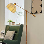Close angle on the pleated shade and brass counterweight of this Clérachic floor lamp, above a green armchair and a small plant.