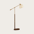 Walnut colour version of this Clérachic floor lamp on a plain warm-grey backdrop, its pleated beige shade held on a brass arm.
