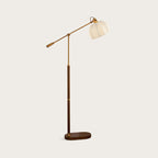 Walnut colour version of this Clérachic floor lamp on a plain warm-grey backdrop, its pleated beige shade held on a brass arm.