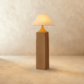 Clérachic floor lamp with a square raw-wood post, a tapered amber-orange neck and a wide linen cone shade glowing warm on limewash.