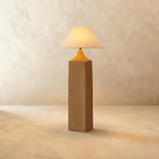 Clérachic floor lamp with a square raw-wood post, a tapered amber-orange neck and a wide linen cone shade glowing warm on limewash.