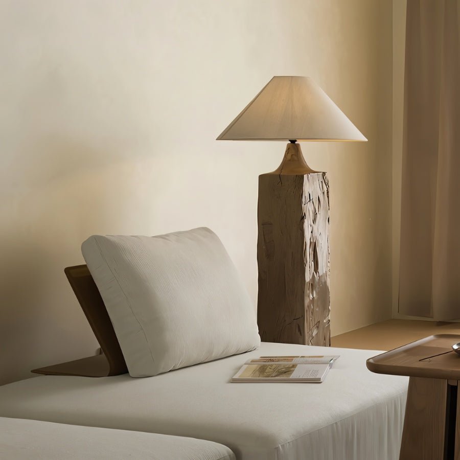 Clérachic floor lamp with a rough-hewn square wooden post and linen cone shade, behind a cream daybed with a pillow and an open magazine.