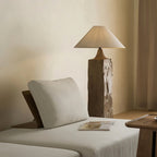 Clérachic floor lamp with a rough-hewn square wooden post and linen cone shade, behind a cream daybed with a pillow and an open magazine.
