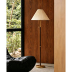 Beside a dark wood-panelled wall and a tall window, this Clérachic floor lamp stands unlit with its pleated beige shade above a black leather chair.