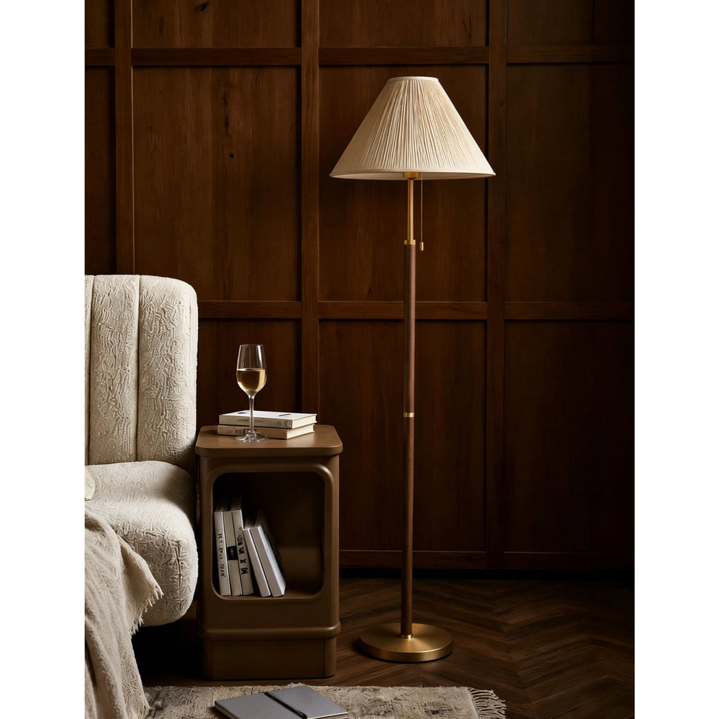 A Clérachic floor lamp with a pleated cream shade beside a cream bouclé armchair and a small side table holding books and a glass of wine.
