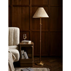 A Clérachic floor lamp with a pleated cream shade beside a cream bouclé armchair and a small side table holding books and a glass of wine.