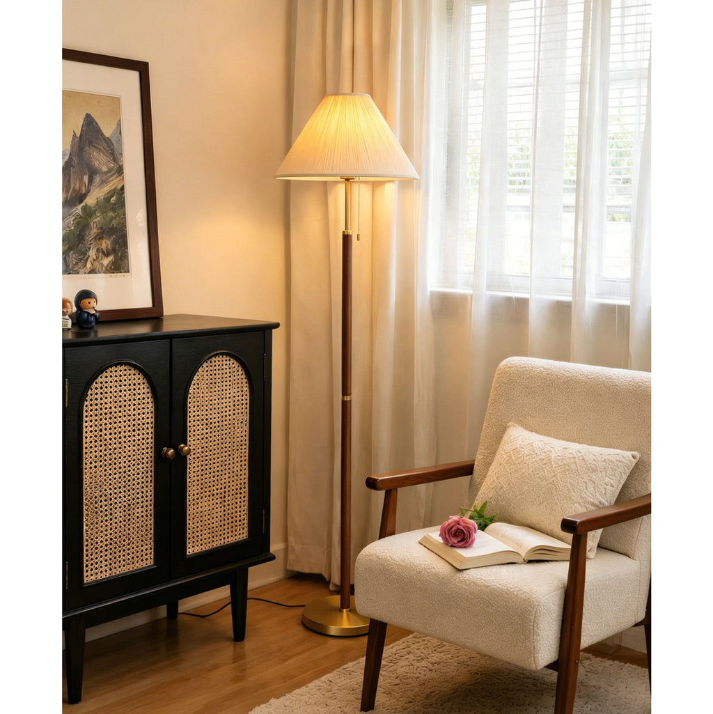 Lit warmly beside a sheer-curtained window, this Clérachic floor lamp stands near a cane-doored black cabinet and an armchair with an open book.