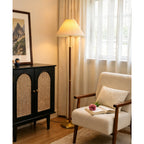 Lit warmly beside a sheer-curtained window, this Clérachic floor lamp stands near a cane-doored black cabinet and an armchair with an open book.