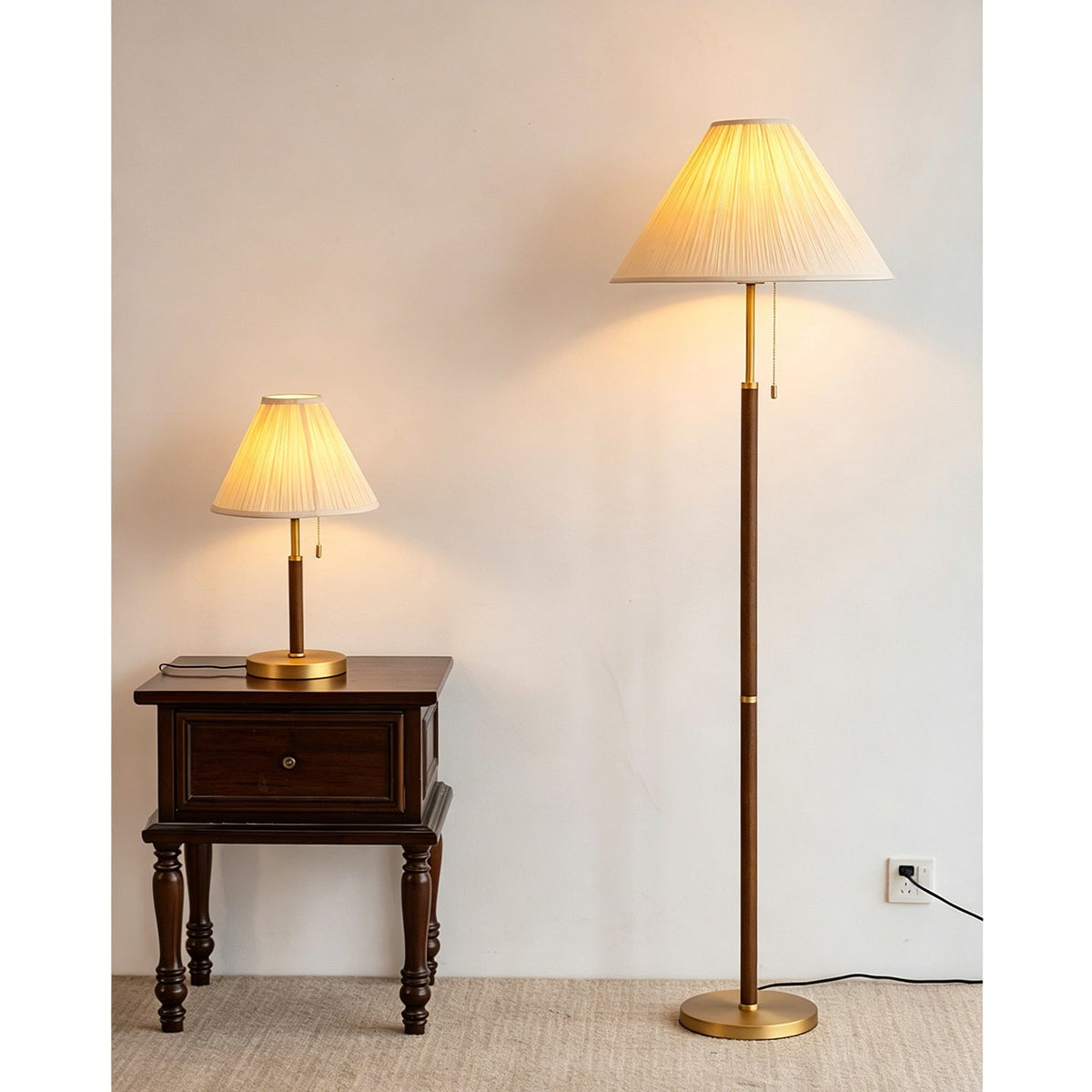 A Clérachic floor lamp lit warm beside a dark carved side table with a small pleated table lamp, on a jute rug against a pale wall.