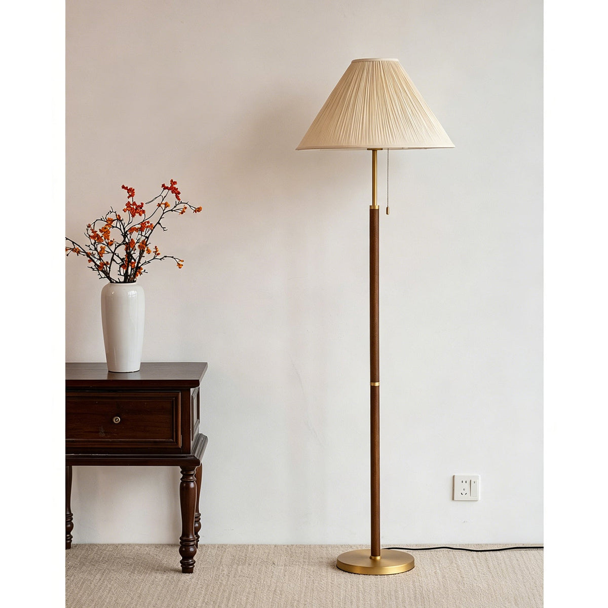 Unlit Clérachic floor lamp with a pleated beige shade, next to a dark side table and a white vase of orange-red berry branches.