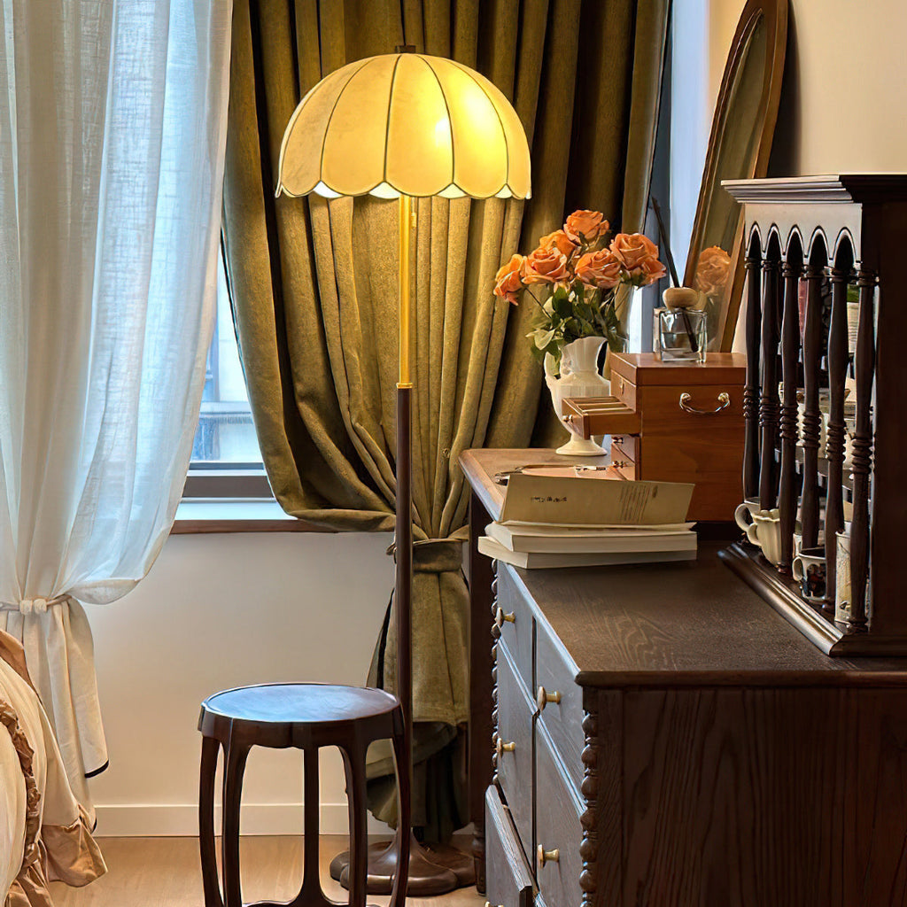 Lit scalloped-shade Clérachic floor lamp beside an olive-green curtain, a dark wooden chest of drawers and a small round stool.
