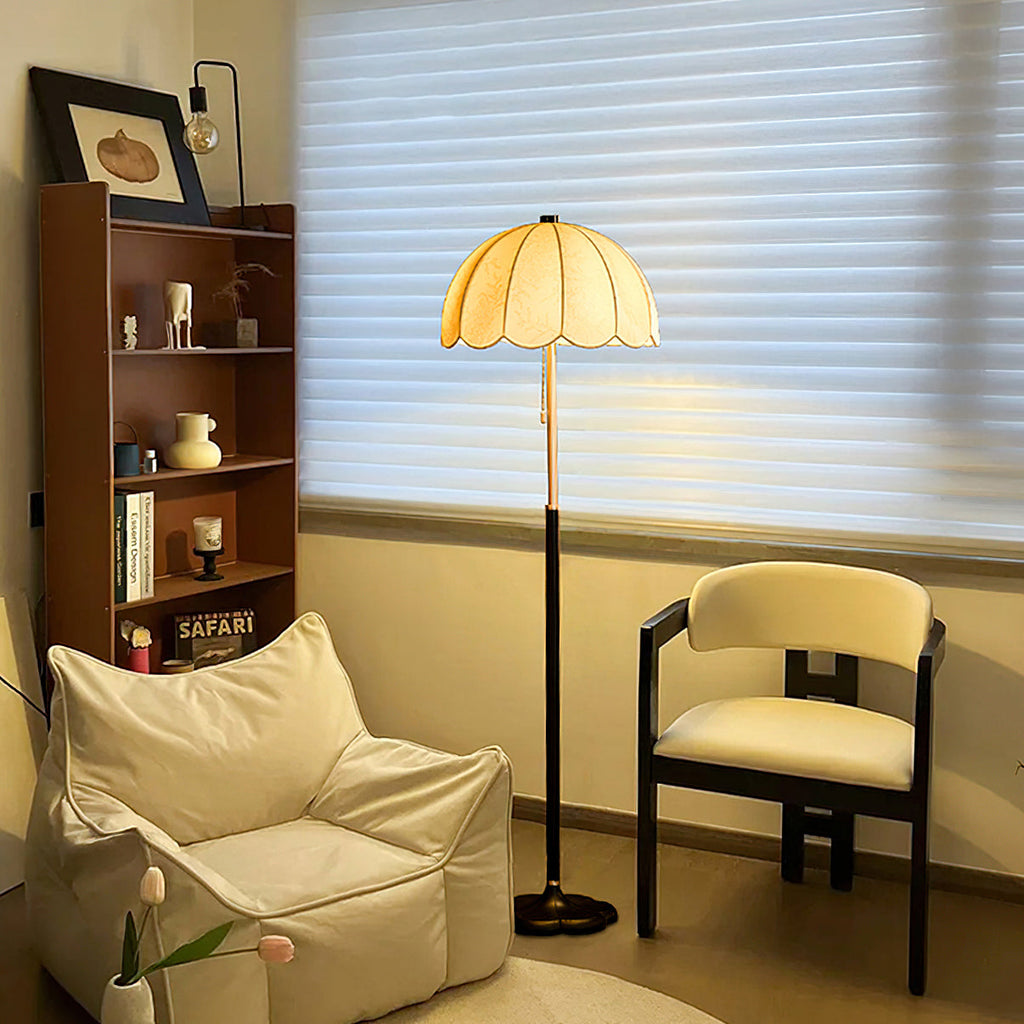 This Clérachic floor lamp glows by a shuttered window, next to a cream armchair, a pale lounge chair and a wooden bookcase.