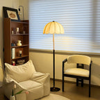 This Clérachic floor lamp glows by a shuttered window, next to a cream armchair, a pale lounge chair and a wooden bookcase.