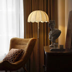Warm evening view of a Clérachic floor lamp beside a tan accent chair and a plaster bust on a dark side table, with a brown curtain behind.