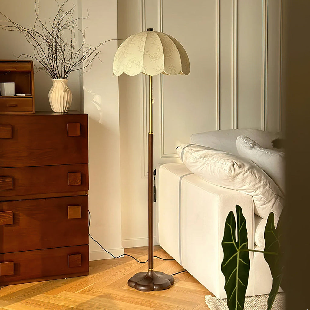 Clérachic floor lamp in a bedroom corner, beside a tall walnut chest of drawers, a white bench with pillows and a large green leaf.