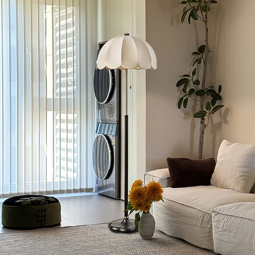 Pale scalloped shade of this Clérachic floor lamp seen in a bright room with vertical blinds, a white sofa and a vase of yellow flowers.