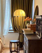 Close-framed view of a lit Clérachic floor lamp shade between a pale blue curtain and an olive-green one, above a dark wooden chest.