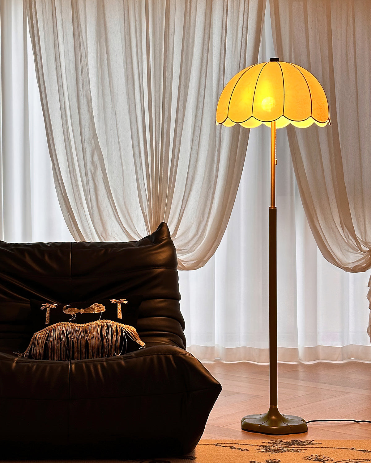 Deep amber glow from the scalloped shade of this Clérachic floor lamp, set before sheer curtains and a dark leather armchair.