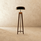 Clérachic tall tripod floor lamp in dark stained wood with a black disc shade, lit, on a limewash backdrop.