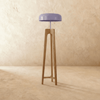 Clérachic tall light wood tripod floor lamp with a lilac disc shade, switched off, standing on a plaster-toned floor.