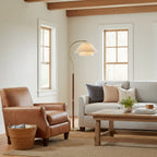 A Clérachic arched floor lamp beside a brown leather armchair and grey sofa in a bright room with timber beams.