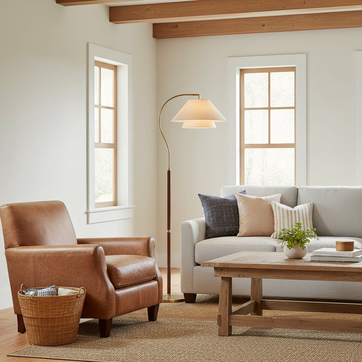 A Clérachic arched floor lamp beside a brown leather armchair and grey sofa in a bright room with timber beams.