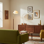A Clérachic floor lamp stands behind an olive sofa, beside a walnut sideboard hung with three small geometric prints.