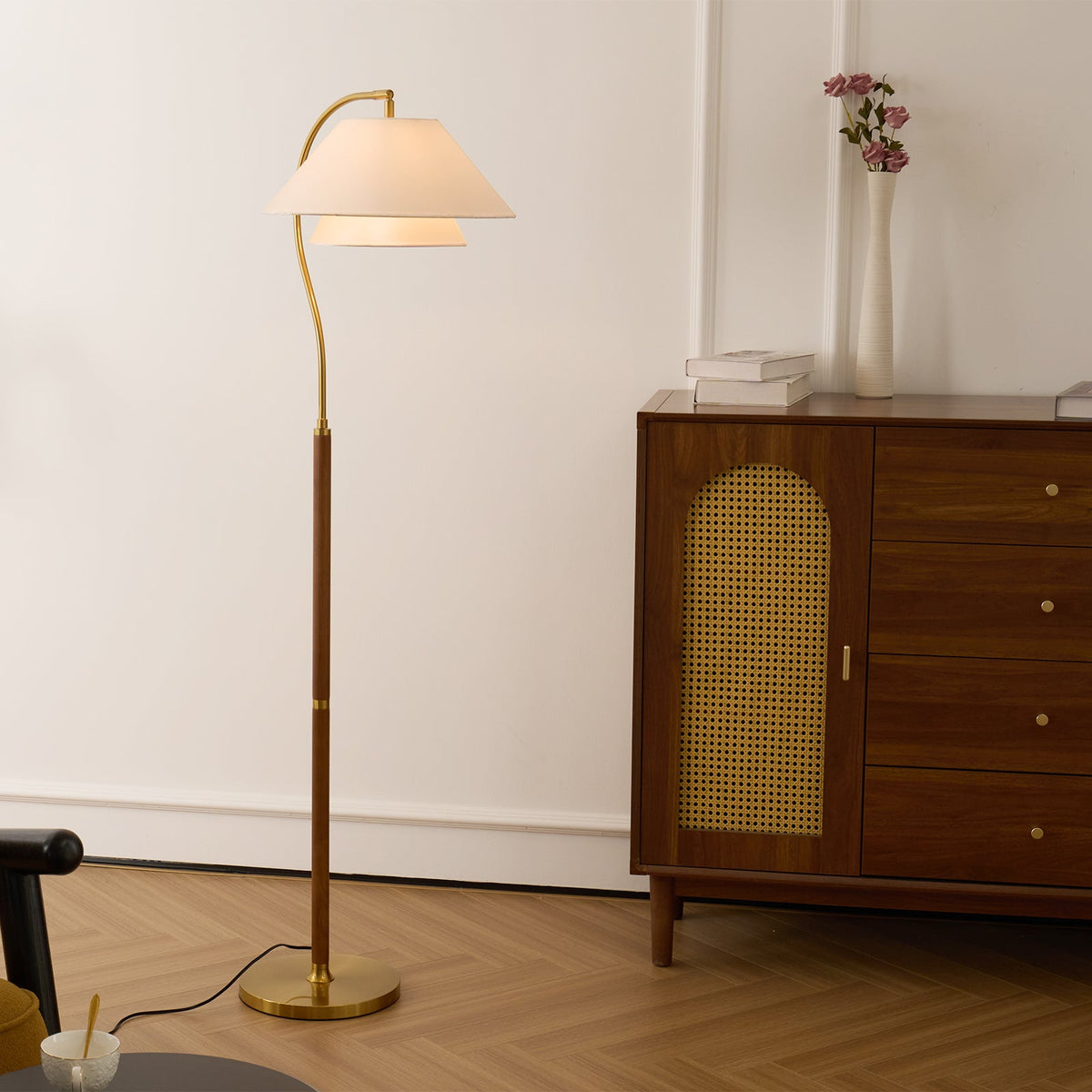 Slim Clérachic floor lamp on a herringbone floor, next to a walnut cabinet with a cane-panel door and a vase of pink flowers.
