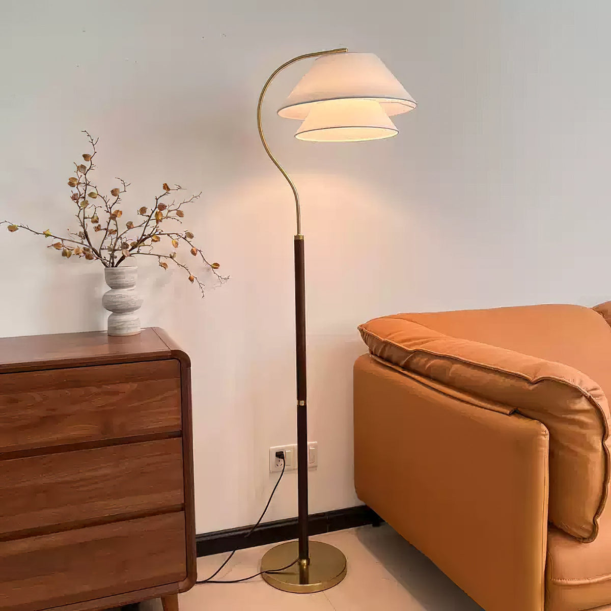 A Clérachic floor lamp with a two-tier shade beside an orange leather sofa and a walnut chest, its cord running to a wall socket.