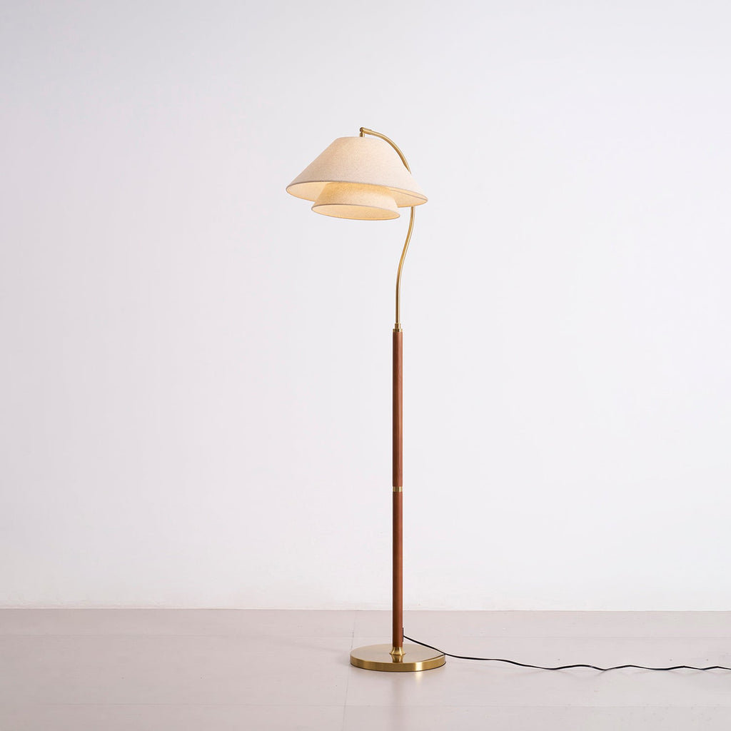 The Clérachic floor lamp switched on, standing on a grey studio floor with its brass base and curved arm in view.