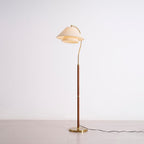The Clérachic floor lamp switched on, standing on a grey studio floor with its brass base and curved arm in view.