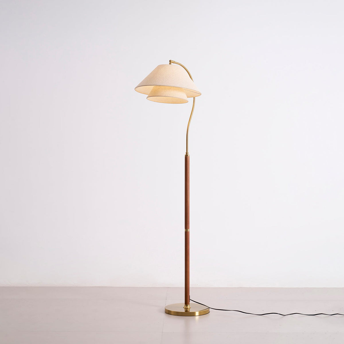 The Clérachic floor lamp switched on, standing on a grey studio floor with its brass base and curved arm in view.
