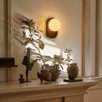 Clérachic wall lamp glowing above a mantel styled with small sculptures and a leafy branch.