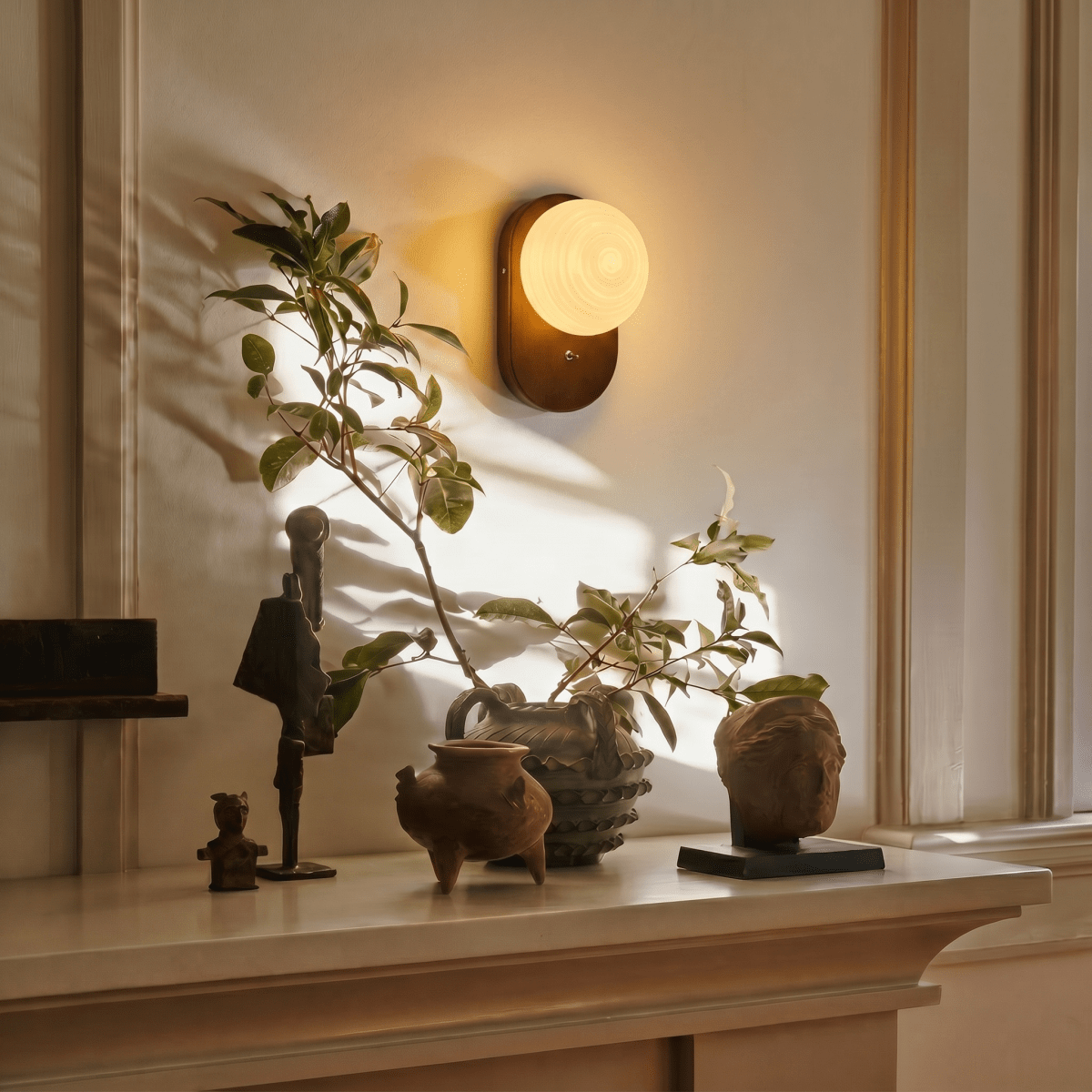 Clérachic wall lamp glowing above a mantel styled with small sculptures and a leafy branch.