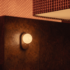 Clérachic wall lamp glowing on a dark wood-paneled bedroom wall beside patterned bedding and a folded throw.