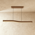 A Clérachic wood-grain bar pendant curves in a gentle wave, lit along its length against a plastered ceiling corner.