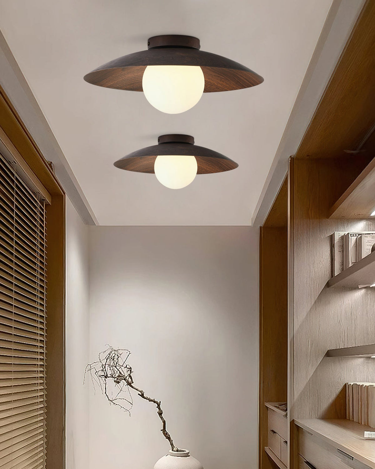 A pair of Clérachic walnut-toned ceiling lights run down a corridor ceiling, with wood shelves on one side and a bare branch in a vase.