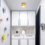 A yellow Clérachic Wood-Grain Cylinder Ceiling Light throwing a cone of light down a narrow entry over white cabinets.