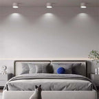 Three white Clérachic Wood-Grain Cylinder Ceiling Lights spaced above a grey upholstered bed, each throwing a tight cone.