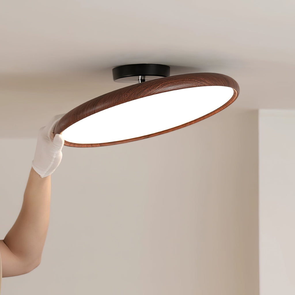 A gloved hand tilts the dark walnut-grain ring of this Clérachic ceiling light to show its white diffuser, below a black ceiling canopy.