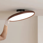A gloved hand tilts the dark walnut-grain ring of this Clérachic ceiling light to show its white diffuser, below a black ceiling canopy.