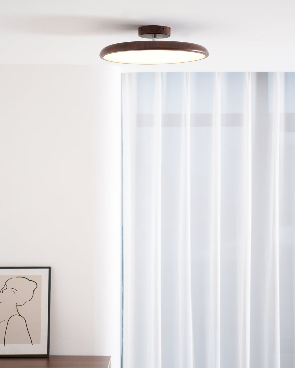 A walnut-grain Clérachic ceiling light above sheer white curtains and a framed line drawing of a figure.
