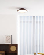 This Clérachic ceiling light in walnut grain, hung above a sideboard with a cane panel and white curtains behind it.