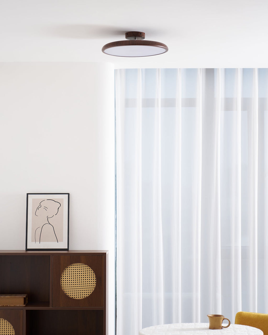 Walnut-grain Clérachic ceiling light seen from a distance above a dark sideboard, a framed drawing and sheer curtains.