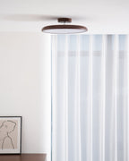 A Clérachic ceiling light with a walnut-grain ring above a white room with sheer curtains and a framed portrait sketch.