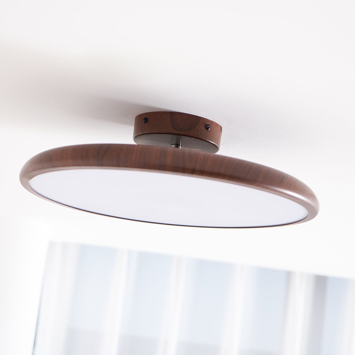 Angled view of this Clérachic ceiling light in walnut grain, with its white diffuser and a sheer curtain below.
