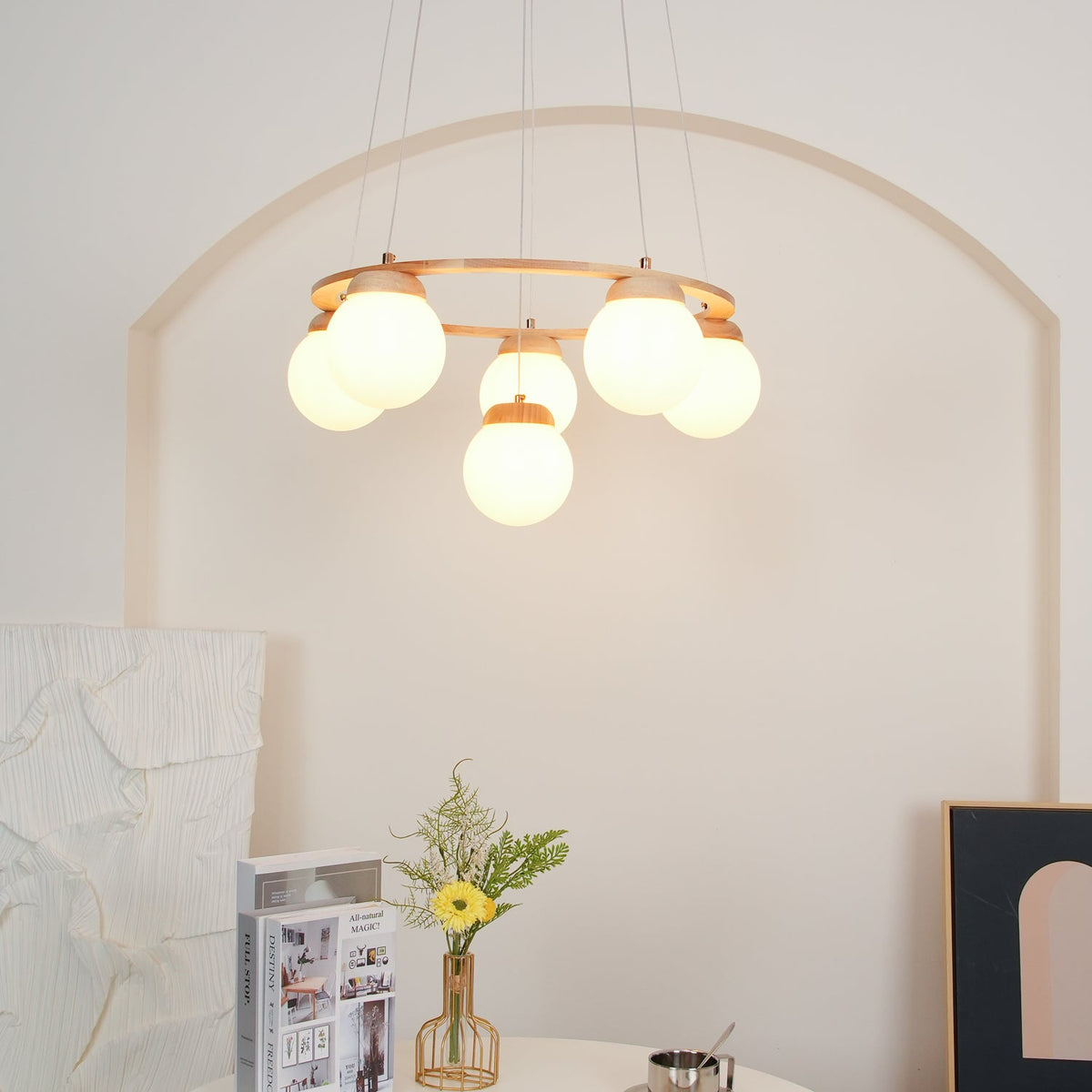 A Clérachic chandelier with frosted globes glowing under a wood ring, above a white table with flowers and magazines.