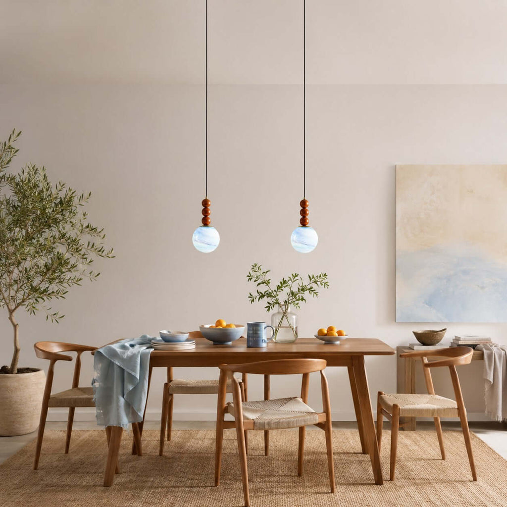 Two Clérachic pendants with pale blue globes and wood stems hang over a timber dining table, with a soft abstract painting behind.